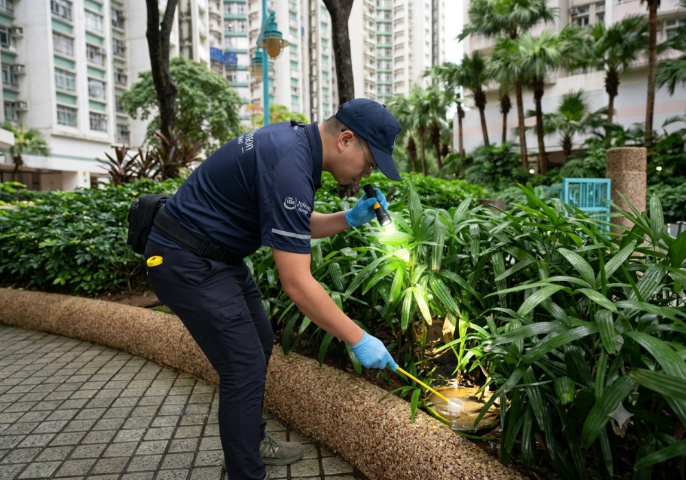Johnson_Group_Mosquitoes_Control_蚊患勘察與場地檢查 身穿深藍制服與手套的防治人員,在住宅屋苑花槽上方,以手電筒照亮積水,並用連接長柄的白色取樣杯檢查是否有蚊蟲孳生。